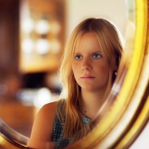 Jeune femme aux cheveux blonds réfléchissant à son reflet dans un miroir doré, symbolisant l'acceptation de soi après une transformation esthétique telle qu'une chirurgie maxillo-faciale.